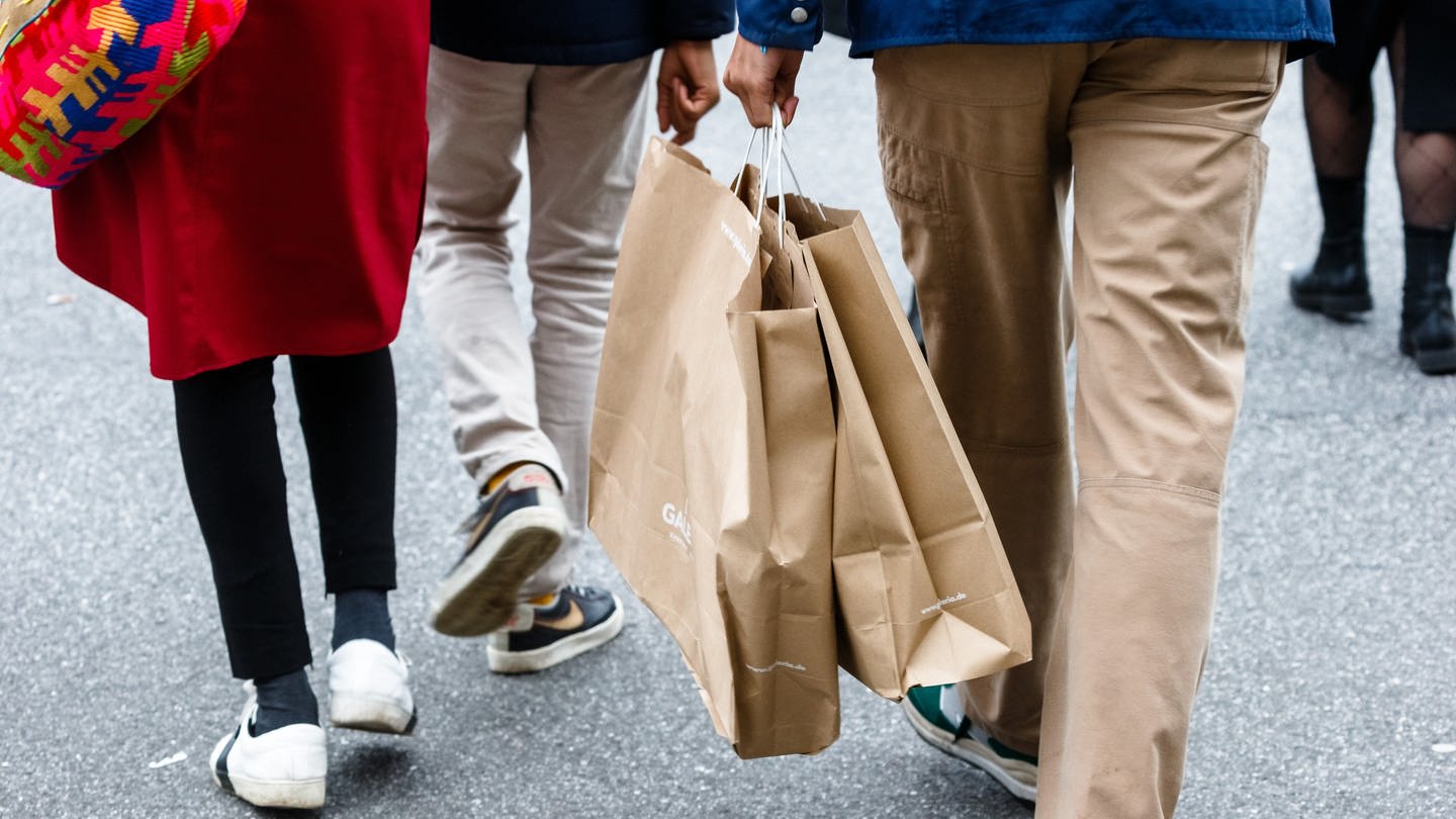 Beine von Menschen. Ein Mensch hält zwei Shoppingtüten in der Hand.