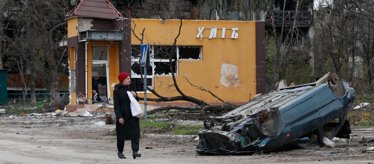 Eine Frau geht in einer ukrainischen Stadt eine Straße entlang und an einem zerstörten auf dem Dach liegenden Auto vorbei. (Foto: dpa Bildfunk, picture alliance/dpa/XinHua | Victor)