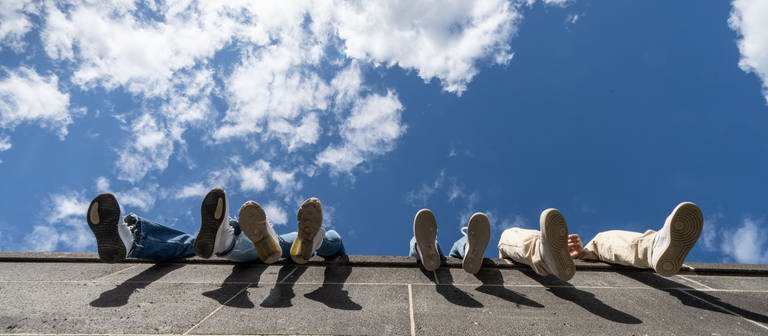 Vier Jugendliche lassen auf einer hohen Mauer am Hafenpark ihre Beine vor dem locker bewölkten Himmel baumeln. (Foto: dpa Bildfunk, picture alliance/dpa | Frank Rumpenhorst)