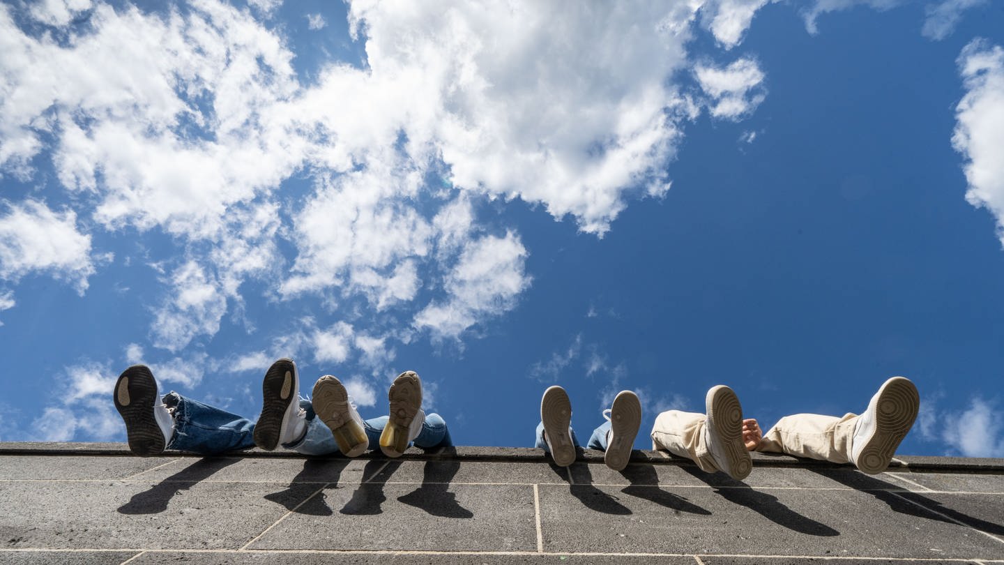Vier Jugendliche lassen auf einer hohen Mauer am Hafenpark ihre Beine vor dem locker bewölkten Himmel baumeln.