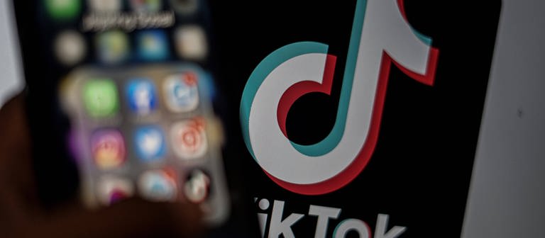 Das TikTok-Logo wird auf einem Monitor im Hintergrund angezeigt, davor wird ein Smartphone mit verschiedenen App-Logis gehalten. (Foto: IMAGO, IMAGO / ZUMA Wire)
