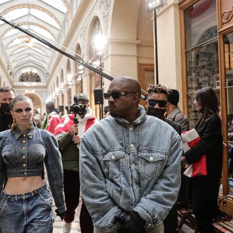 Kanye West in Jeansjacke und mit Sonnenbrille, gefolgt von seiner Freundin und Kameraleuten