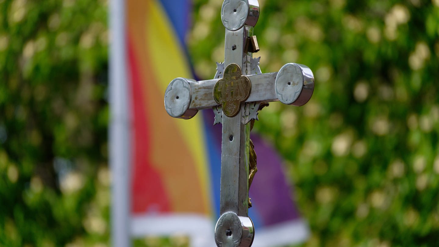 Ein Kreuz steht bei einem Open-Air-Segnungsgottesdienst für Liebende vor der Kirche Christi Auferstehung. Im Hintergrund ist eine Regenbogenfahne zu sehen.