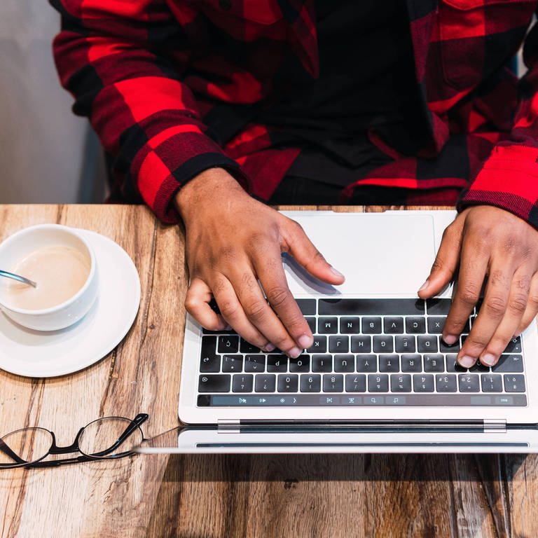 Draufsicht auf einen Tisch mit Kaffee, Notebook und Händen auf der Tastatur. (Foto: IMAGO, IMAGO / Addictive Stock)