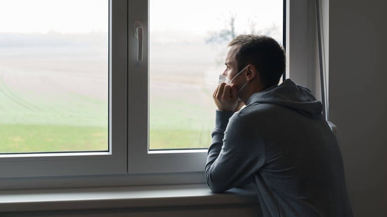 Ein junger Mann steht mit FFP2-Maske an einem Fenster und schaut nach draußen.