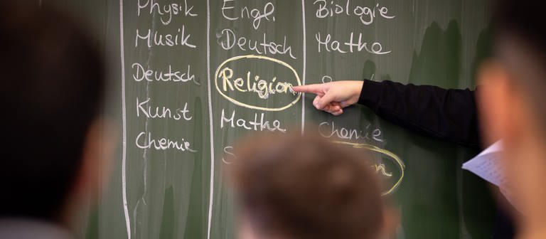 Schülerin Delin steht an einer Tafel, auf der ein Stundenplan mit Religionsunterricht geschrieben ist. (Foto: dpa Bildfunk, picture alliance/dpa | Friso Gentsch)