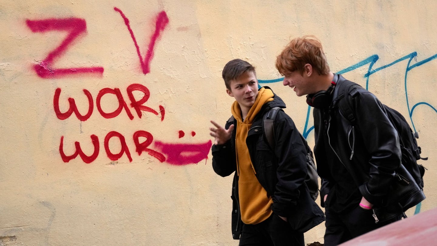 Zwei junge Männer gehen an einer Wand mit einem Graffiti vorbei.