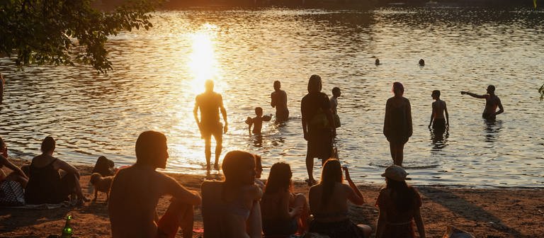 An einem See sitzen am Abend während des Sonnenuntergangs einige junge Menschen, andere sind im Wasser. (Foto: dpa Bildfunk, picture alliance/dpa | Jörg Carstensen)