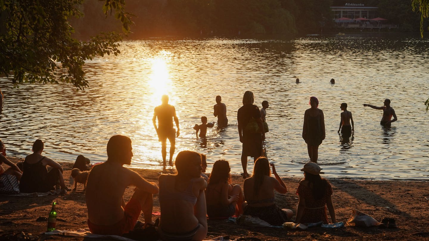 An einem See sitzen am Abend während des Sonnenuntergangs einige junge Menschen, andere sind im Wasser.