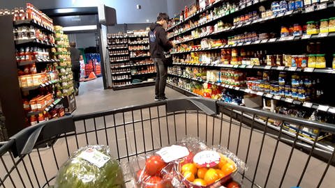 Ein junger Mann steht in einem Supermarkt an einem Regal mit Lebensmittel, im Vordergrund ist ein Einkaufswagen zu sehen mit Obst und Gemüse. (Foto: IMAGO, IMAGO / Martin Wagner)
