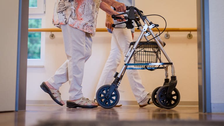 Eine Pflegekraft geht in einem Pflegeheim mit einer älteren Dame über einen Korridor.  (Foto: dpa Bildfunk, picture alliance/dpa | Christoph Schmidt)