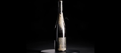 Eine Weinflasche aus der Schlammflut in Ahrtal steht auf einem schwarzen Ständer (Foto: DASDING, Screenshot: https://www.niftee.eu/de/title/FLUTWEIN-14)