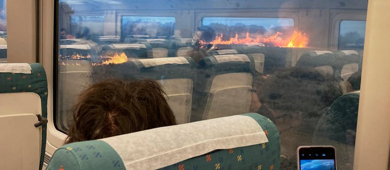 Fahrgäste fotografieren während einer Zugfahrt einen Flächenbrand. Der Zug hielt unterwartet in einer Region an, in der sich die Flammen ausbreiteten. Fahrgäste sollen voller Angst beobachtet haben, wie schnell sich Flammen ausbreiten. In Spanien waren am Montag noch 22 Feuer aktiv. (Foto: dpa Bildfunk, picture alliance/dpa/AP | Francisco Seoane Perez)