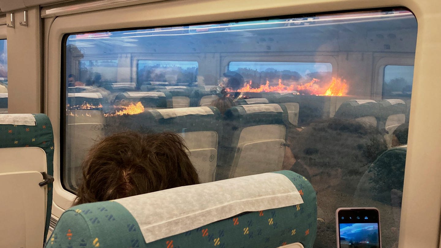 Fahrgäste fotografieren während einer Zugfahrt einen Flächenbrand. Der Zug hielt unterwartet in einer Region an, in der sich die Flammen ausbreiteten. Fahrgäste sollen voller Angst beobachtet haben, wie schnell sich Flammen ausbreiten. In Spanien waren am Montag noch 22 Feuer aktiv.