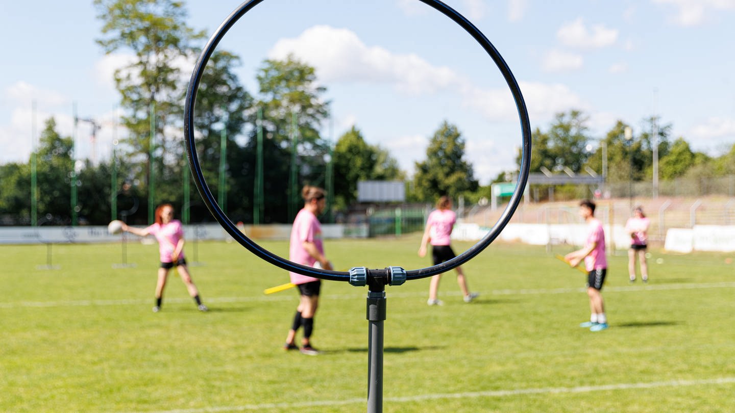 Ein Ständer mit einem Ring, der als Tor dient, steht auf dem Spielfeld. Quidditch ist ein Kontaktsport, der in gemischtgeschlechtlichen Teams gespielt wird und Elemente aus verschiedenen Ballsportarten miteinander vereint.