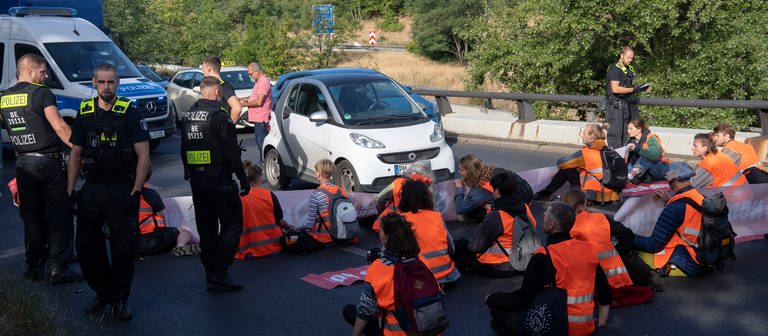 Aktivisten der Gruppe "letzte Generation" sitzen auf einer Straße und blockieren so den Verkehr. Um sie herum stehen mehrere Polizisten. (Foto: dpa Bildfunk, picture alliance/dpa | Paul Zinken)