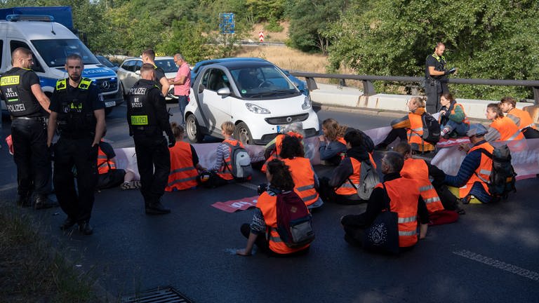 Aktivisten der Gruppe "letzte Generation" sitzen auf einer Straße und blockieren so den Verkehr. Um sie herum stehen mehrere Polizisten. (Foto: dpa Bildfunk, picture alliance/dpa | Paul Zinken)