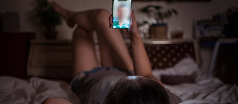 Eine junge Frau liegt aut einem Bett und schaut auf ihr Handy, auf dessen Display ist das Portrait eines jungen Mannes zu sehen. (Foto: IMAGO, IMAGO / Addictive Stock)