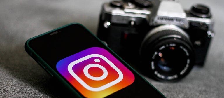 Auf einem Handydisplay wird groß das Instagram-Logo angezeigt. Daneben liegt eine Foto-Kamera. (Foto: IMAGO, IMAGO / NurPhoto)