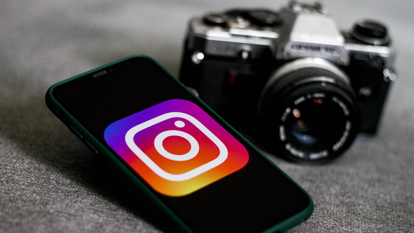Auf einem Handydisplay wird groß das Instagram-Logo angezeigt. Daneben liegt eine Foto-Kamera. (Foto: IMAGO, IMAGO / NurPhoto)