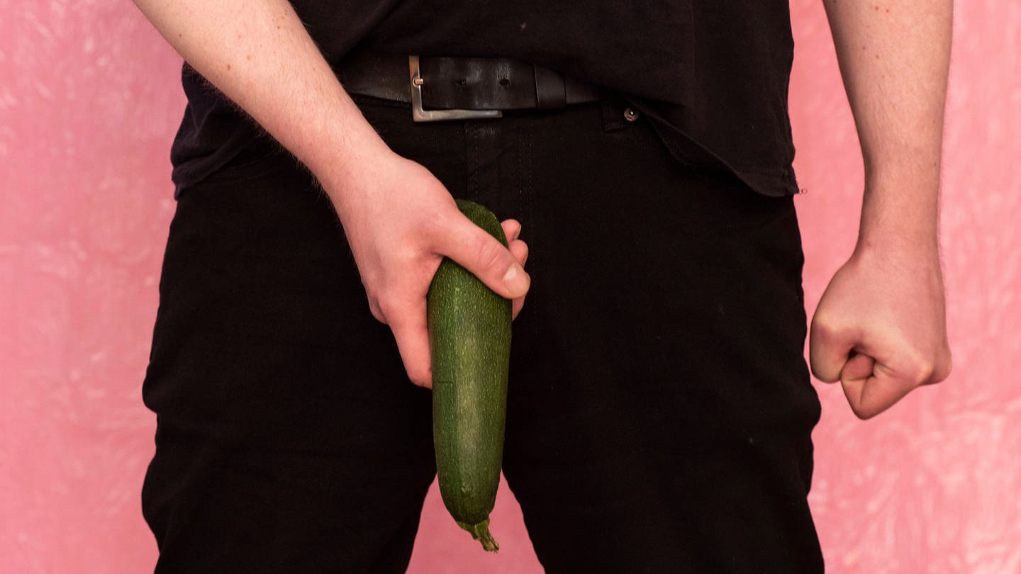 Ein Mann hält eine Zucchini als Penis an seine Hose