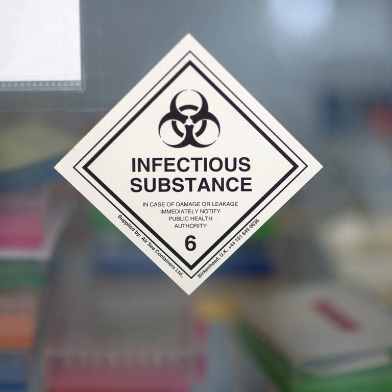 Auf einer Glastür, hinter der man verschiedenen Aufbewahungsboxen erkennen kann, steht auf einem Hinweis-Aufkleber "Infectious Substance". (Foto: IMAGO, IMAGO / IPON)