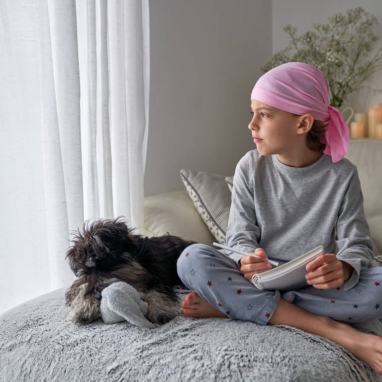  little child with cancer disease writing notes while sitting with dog on bed in room  (Foto: IMAGO, IMAGO / Addictive Stock;  Copyright: xPhilippexDegrootex)