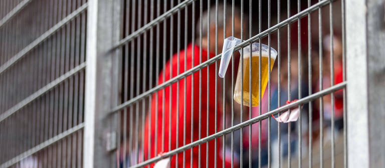 Ein Becher mit Bier hängt im Stadionzaun. (Foto: IMAGO, Sascha Janne)