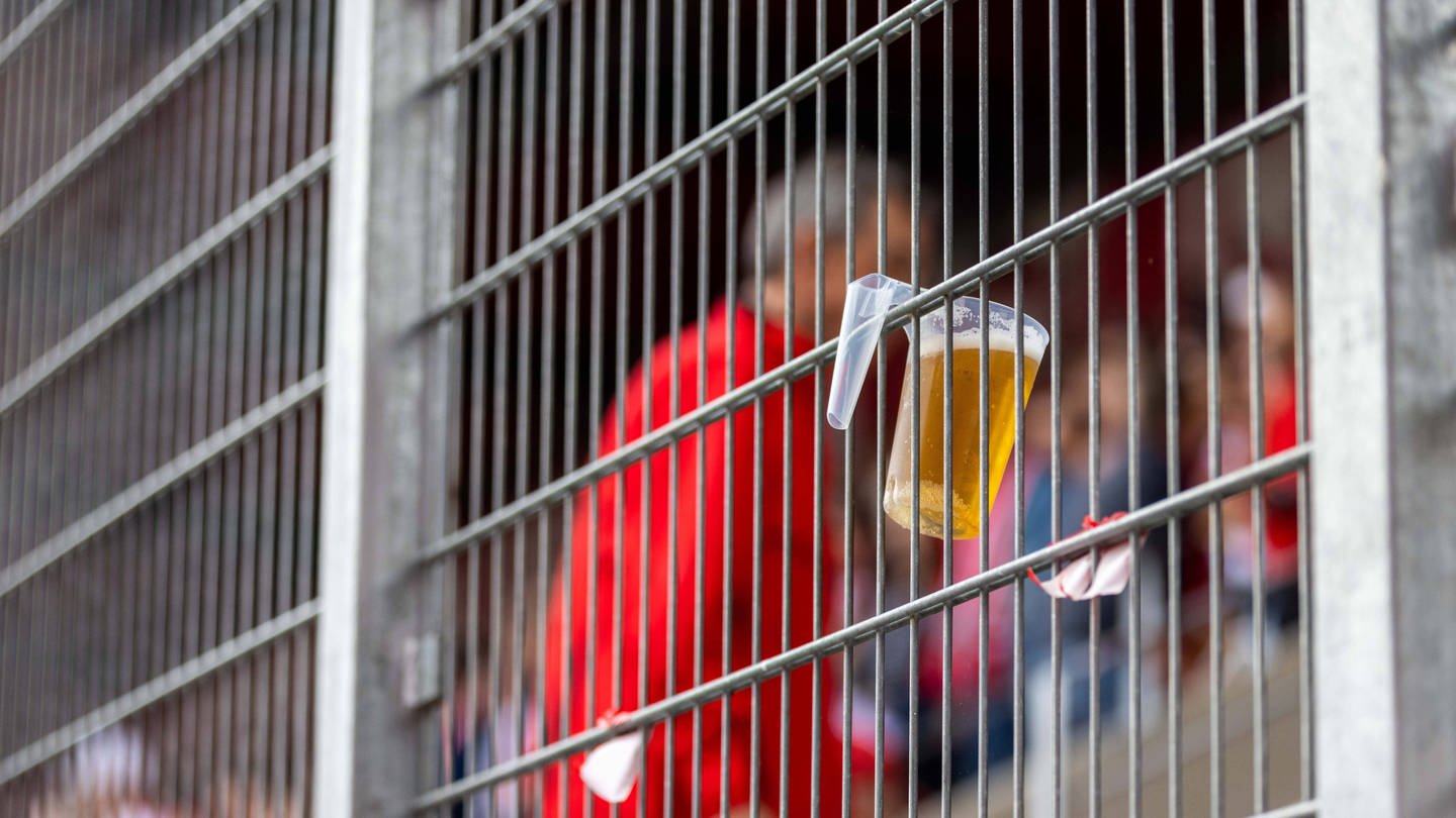 Ein Becher mit Bier hängt im Stadionzaun.
