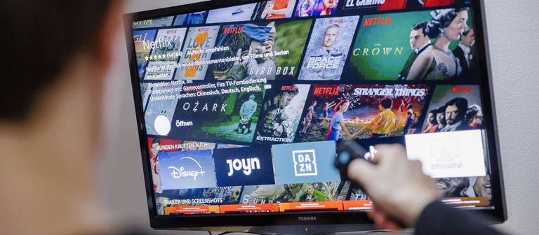 Auf einem Fernseher sind verschiedene Logos von Streaming-Anbietern zu sehen. Ein junger Mann sitzt davor und zeigt mit der Fernbedienung auf den TV. (Foto: IMAGO, IMAGO / photothek)