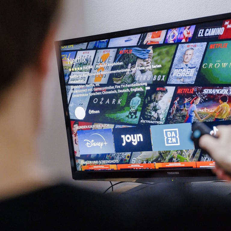Auf einem Fernseher sind verschiedene Logos von Streaming-Anbietern zu sehen. Ein junger Mann sitzt davor und zeigt mit der Fernbedienung auf den TV. (Foto: IMAGO, IMAGO / photothek)
