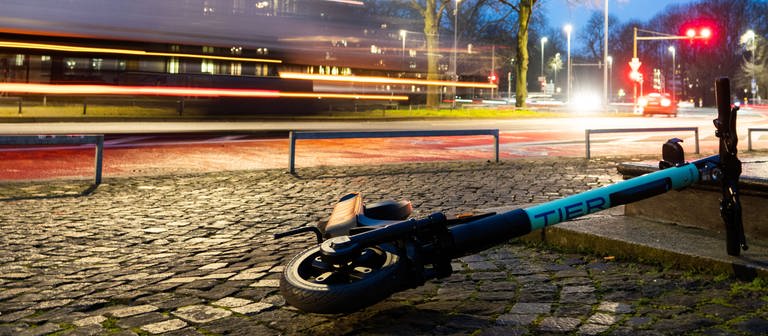 Ein E-Scooter liegt in der Morgendämmerung auf einem Gehweg in der Innenstadt, während auf der Straße LKW vorbeifahren. (Foto: dpa Bildfunk, picture alliance/dpa | David Hutzler)