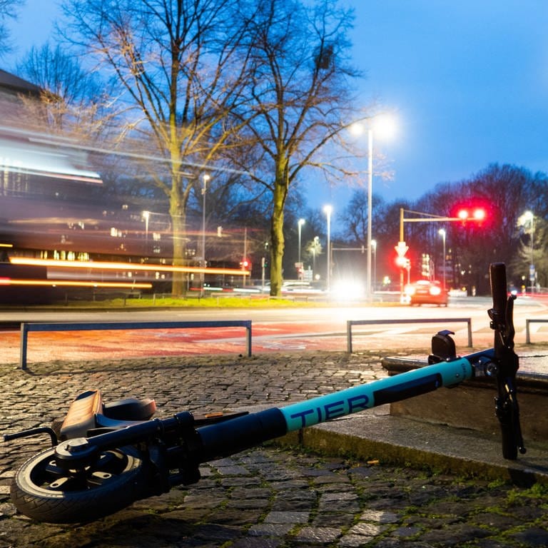 Ein E-Scooter liegt in der Morgendämmerung auf einem Gehweg in der Innenstadt, während auf der Straße LKW vorbeifahren. (Foto: dpa Bildfunk, picture alliance/dpa | David Hutzler)