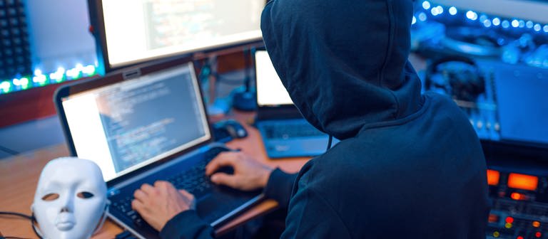 Hacker in mask and hood, account hacking, Hacker in mask and hood sitting at his workplace with laptop and PC, password or account hacking. (Foto: IMAGO, IMAGO / Panthermedia)
