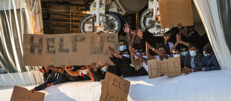 Migranten halten Transparente mit der Bitte um Hilfe an Deck des unter norwegischer Flagge fahrenden Schiffes «Geo Barents», das von «Ärzte ohne Grenzen» betrieben wird, im Hafen von Catania (Foto: dpa Bildfunk, picture alliance/dpa/AP | Salvatore Cavalli)