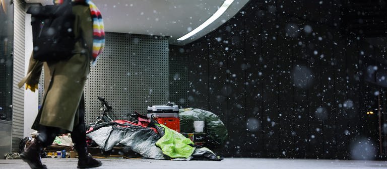 ine Frau geht am frühen Morgen an einem Schlafplatz von obdachlosen Menschen unter dem Vordach eines Kaufhauses in der Innenstadt vorbei.  (Foto: dpa Bildfunk, picture alliance/dpa | Christian Charisius)