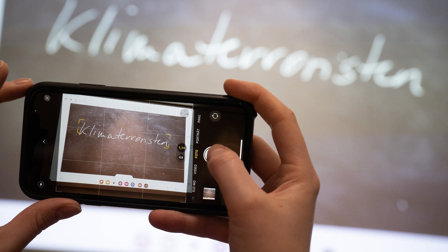 Eine Journalistin fotografiert mit ihrem Smartphone das «Unwort des Jahres» 2022, «Klimaterroristen», das während seiner Bekanntgabe an der Philipps-Universität an eine Wand projiziert wird. (Foto: dpa Bildfunk, picture alliance/dpa | Sebastian Gollnow)