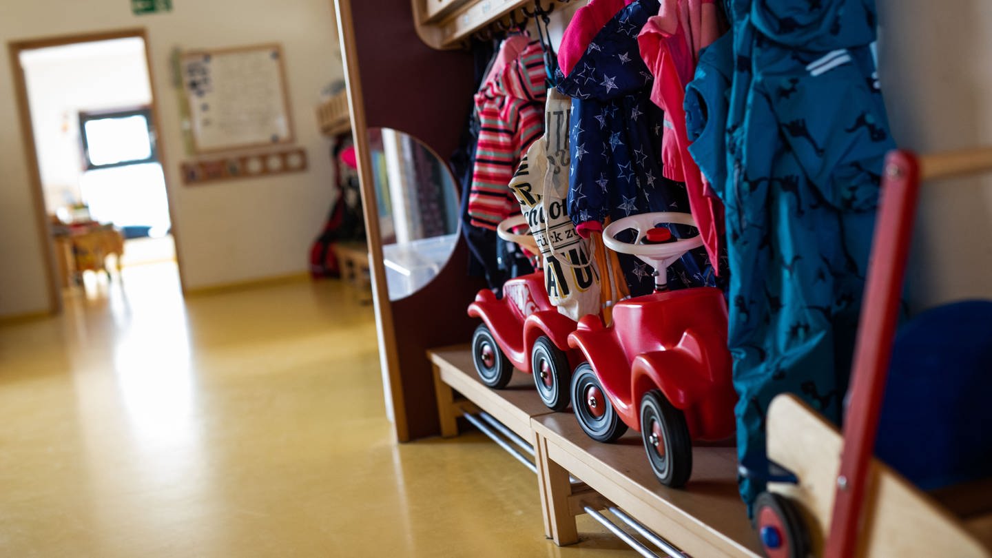 Bobbycars stehen unter Jacken von Kindern, die an einer Garderobe einer Kindertagesstätte (Kita) hängen. (Foto: dpa Bildfunk, picture alliance/dpa | Philipp von Ditfurth)