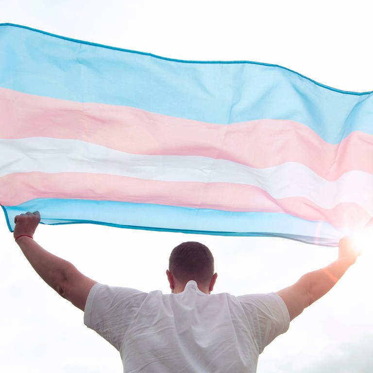 Ein junger Mann hält eine Trans-Pride-Flagge in die Luft (Foto: IMAGO, IMAGO/Pond5)