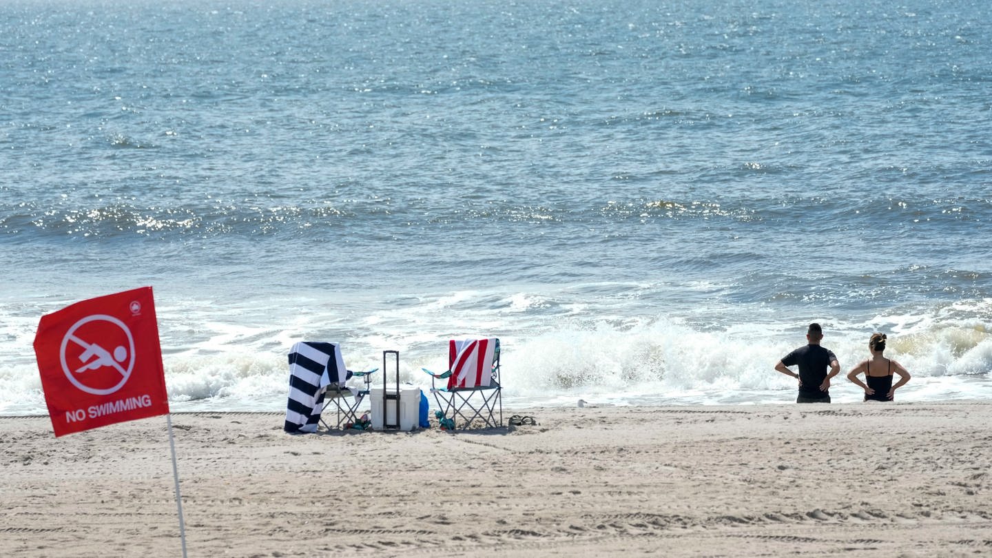 Besucher stehen am Strand vor einer roten Fahne, die für ein Badeverbot am Strand von Rockaway steht. Eine 50-jährige Frau ist an einem Strand der Millionenmetropole New York von einem Hai schwer verletzt worden. (Foto: dpa Bildfunk, picture alliance/dpa/AP | Mary Altaffer)