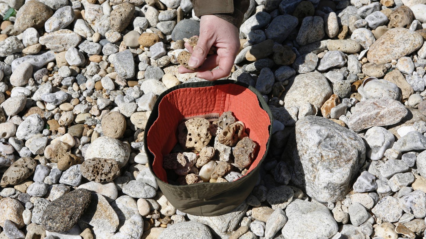 Eine Hand hebt Steine am Strand auf. Einer Touristin aus Belgien droht aktuell eine Haftstrafe, weil sie Steine aus dem Türkei-Urlaub mit nach Hause nehmen wollte. (Foto: IMAGO, robertharding)