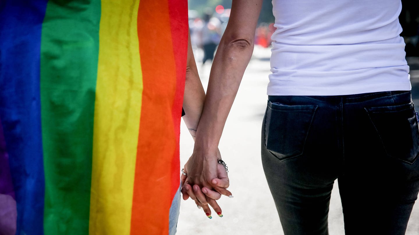 Zwei Personen halten Händchen und haben eine Regenbogen-Flagge umhängen. (Foto: IMAGO, IMAGO / ZUMA Wire)