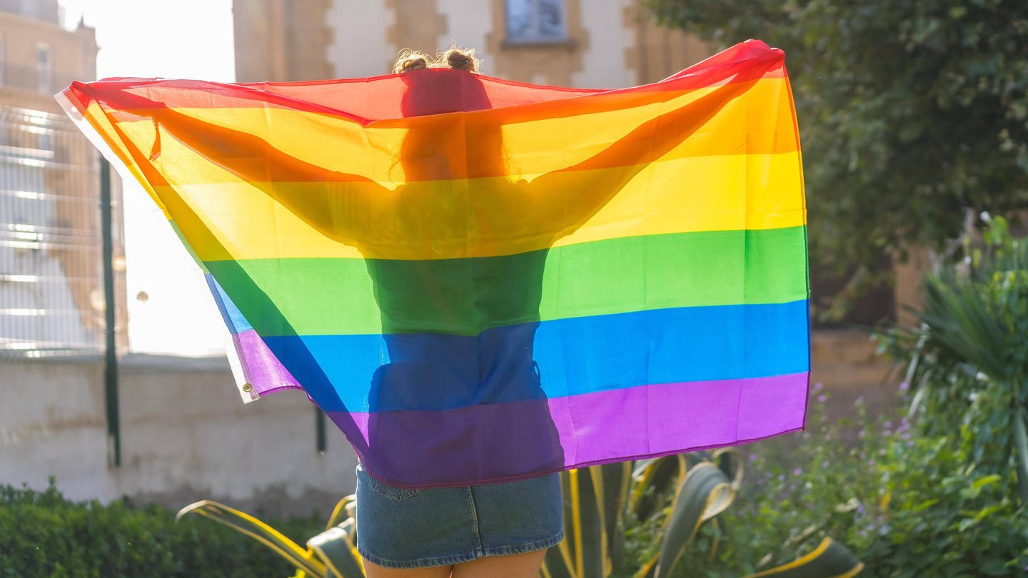 Frau von hinten mit einer Regenbogen-Flagge über den Schultern: In Russland ist die LGBTQ-Community als extremistisch verboten worden. (Foto: IMAGO, IMAGO / imagebroker)