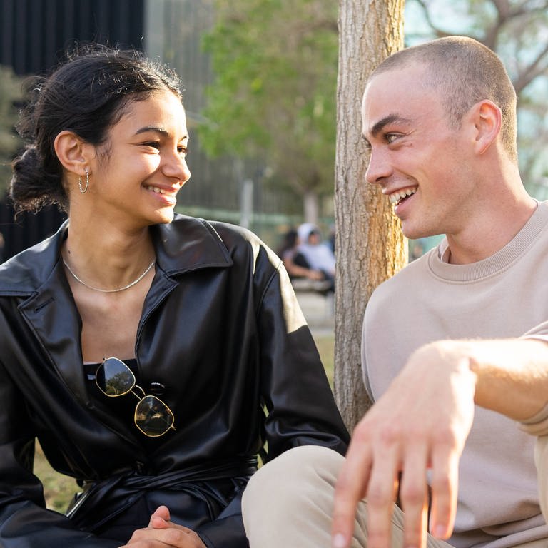 SYMBOLBILD: Eine junge Frau und ein junger Mann sitzen gemeinsam in der Wiese und lachen einander an. (Foto: IMAGO, IMAGO / Addictive Stock)
