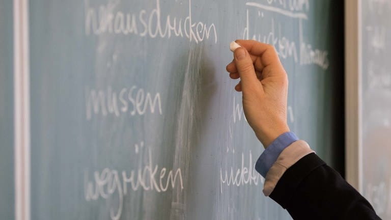 Ein Lehrer steht in einem Klassenraum an einer Tafel und schreibt.