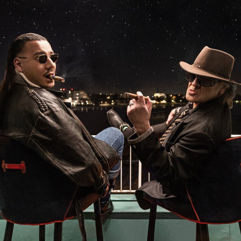 Rapper Apache 207 aus Ludwigshafen und Udo Lindenberg sitzen mit Zigarren nebeneinander auf einem Balkon und blicken in die Nacht (Foto: dpa Bildfunk, picture alliance/dpa/Warner Music | Tine Acke)