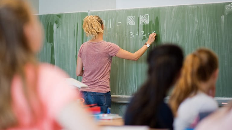 Eine junge Lehrerin schreibt an eine Schultafel: Leherverband fordert mehr Hitzeschutz an Schulen