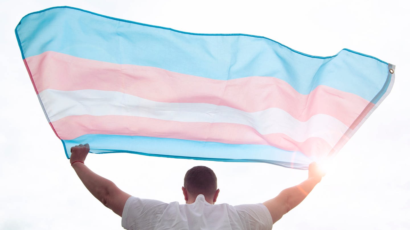 Ein junger Mann hält eine Trans-Pride-Flagge in die Luft (Foto: IMAGO, IMAGO/Pond5)