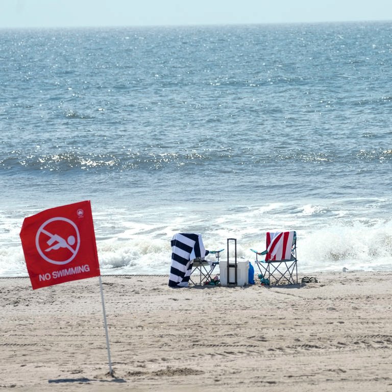 Besucher stehen am Strand vor einer roten Fahne, die für ein Badeverbot am Strand von Rockaway steht. Eine 50-jährige Frau ist an einem Strand der Millionenmetropole New York von einem Hai schwer verletzt worden. (Foto: dpa Bildfunk, picture alliance/dpa/AP | Mary Altaffer)