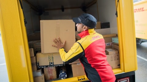 Ein Paketzusteller stellt ein Paket in ein Fahrzeug der Deutschen Post DHL. Es gibt einen neuen Betrugsversuch per Mail. Du wirst aufgefordert eine Zollgebühr für dein Paket zu zahlen. (Foto: dpa Bildfunk, picture alliance / Rolf Vennenbernd/dpa | Rolf Vennenbernd)